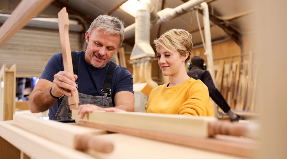 Ein erfahrener Schreiner zeigt einer jungen Frau in einer Werkstatt den Umgang mit einem Holzwerkstück, während sie aufmerksam zusieht. Ein erfahrener Schreiner zeigt einer jungen Frau in einer Werkstatt den Umgang mit einem Holzwerkstück, während sie aufmerksam zusieht.