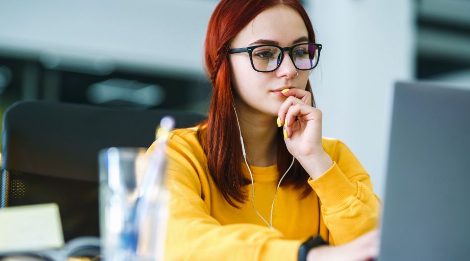 Eine junge Frau mit roten Haaren und Brille sitzt konzentriert vor einem Laptop, trägt gelbe Kleidung und hört über Kopfhörer zu. Eine junge Frau mit roten Haaren und Brille sitzt konzentriert vor einem Laptop, trägt gelbe Kleidung und hört über Kopfhörer zu.