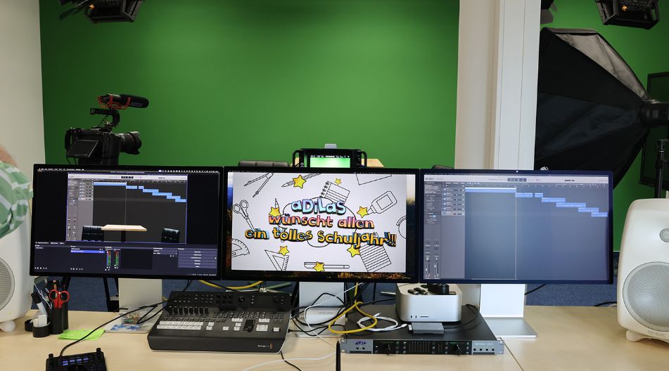 Ein modernes Video- und Tonaufnahmestudio mit drei Monitoren, die eine Videoproduktion und Tonbearbeitung zeigen. Im Hintergrund befindet sich eine grüne Wand (Greenscreen) sowie Kamera- und Beleuchtungsausrüstung. Auf dem mittleren Monitor steht „aDilas wünscht allen ein tolles Schuljahr!!“ in bunten Buchstaben. Ein modernes Video- und Tonaufnahmestudio mit drei Monitoren, die eine Videoproduktion und Tonbearbeitung zeigen. Im Hintergrund befindet sich eine grüne Wand (Greenscreen) sowie Kamera- und Beleuchtungsausrüstung. Auf dem mittleren Monitor steht „aDilas wünscht allen ein tolles Schuljahr!!“ in bunten Buchstaben.