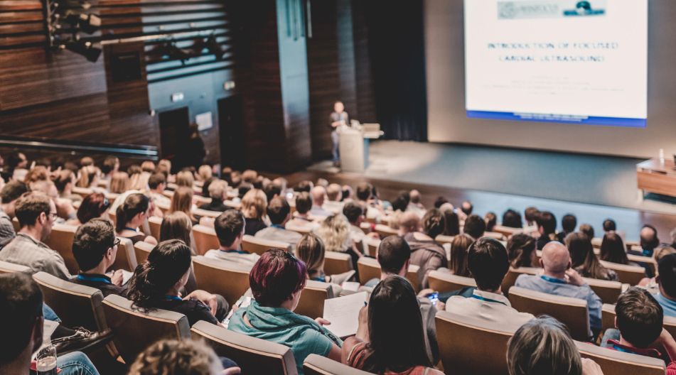 Ein großer Hörsaal mit vielen Zuhörern, die einer Präsentation auf einer Leinwand folgen. Eine Person steht am Rednerpult und hält einen Vortrag. Ein großer Hörsaal mit vielen Zuhörern, die einer Präsentation auf einer Leinwand folgen. Eine Person steht am Rednerpult und hält einen Vortrag.