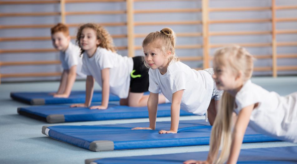 Vier Kinder in Sportkleidung machen gemeinsam eine Gymnastikübung auf blauen Matten in einer Turnhalle. Sie tragen weiße T-Shirts und konzentrieren sich auf die Übung.