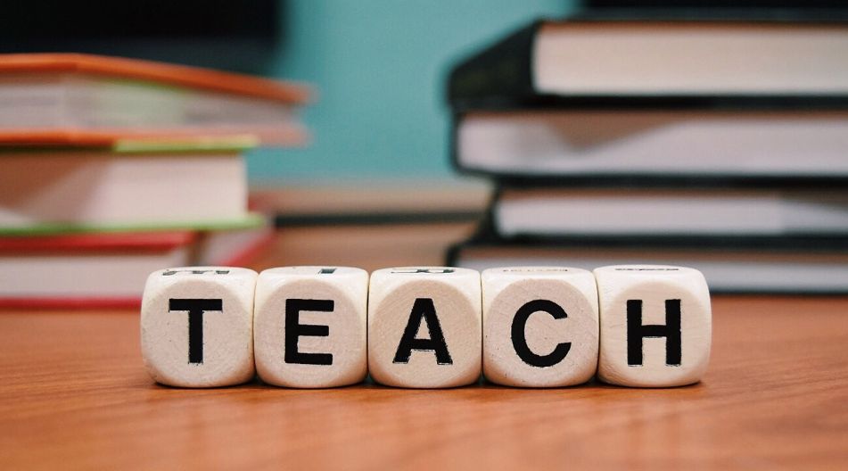 Würfel "Lehren" umgeben von Büchern Würfel mit dem Wort "Lehren" auf einem Schreibtisch, umgeben von Büchern.