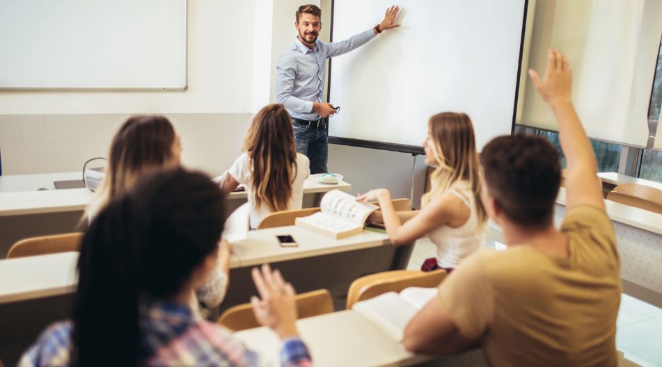 Ein Berufsschullehrer steht vor einer Klasse und zeigt auf eine leere Projektionsfläche, während mehrere Schüler aufmerksam zuhören und einer die Hand hebt.