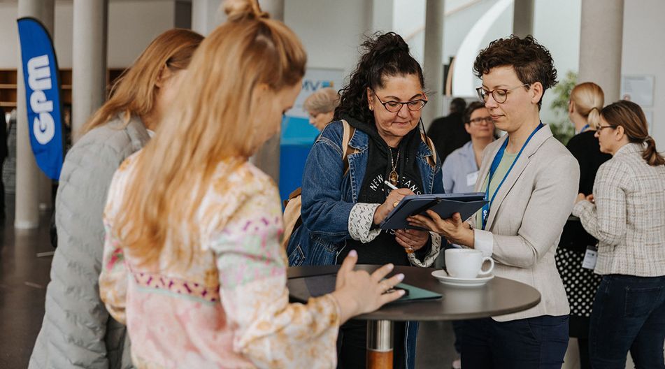 Mehrere Personen stehen bei einer Fachtagung an einem Stehtisch, zwei Frauen betrachten gemeinsam ein Tablet, im Hintergrund sind weitere Teilnehmende im Gespräch.