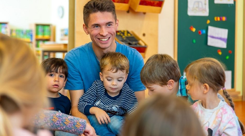 Ein lächelnder Erzieher liest einer Gruppe von Kindern in einem Klassenzimmer ein Buch vor. Ein lächelnder Erzieher liest einer Gruppe von Kindern in einem Klassenzimmer ein Buch vor.