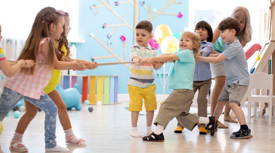 Kinder spielen Tauziehen in einem hellen Raum mit buntem Hintergrund Eine Gruppe von Kindern spielt Tauziehen in einem hellen, farbenfrohen Raum. Die Kinder lächeln und sind in zwei Teams aufgeteilt, die jeweils an einem Seil ziehen, unterstützt von einer Betreuerin im Hintergrund.