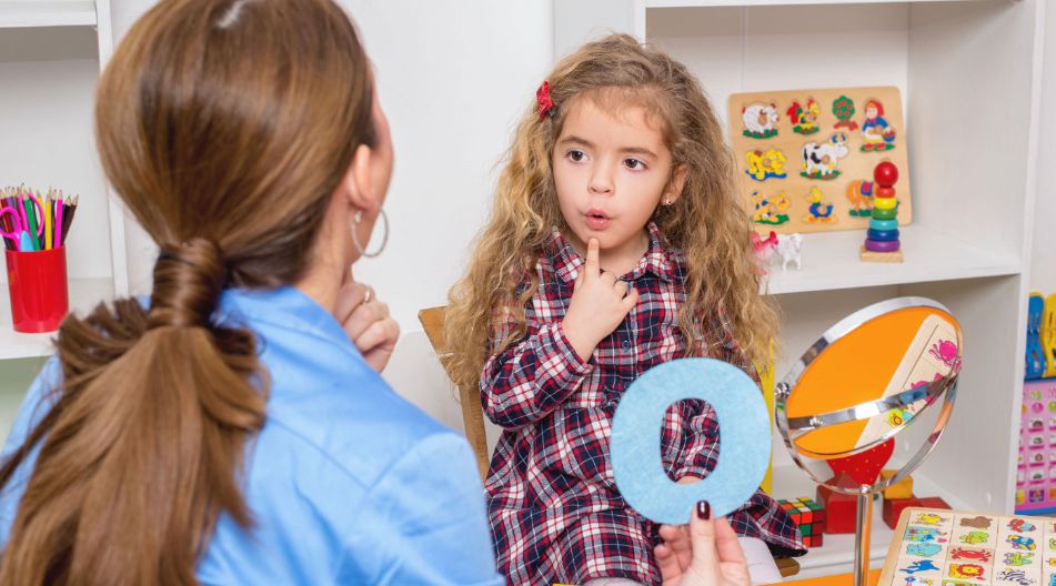 Eine Erzieherin sitzt mit einem jungen Mädchen in einem bunt gestalteten Lernraum. Das Mädchen trägt ein kariertes Kleid und formt mit den Lippen einen Laut, während die Frau einen blauen Buchstaben „O“ hält. Auf dem Tisch liegen Lernmaterialien, und ein Spiegel steht daneben. Eine Erzieherin sitzt mit einem jungen Mädchen in einem bunt gestalteten Lernraum. Das Mädchen trägt ein kariertes Kleid und formt mit den Lippen einen Laut, während die Frau einen blauen Buchstaben „O“ hält. Auf dem Tisch liegen Lernmaterialien, und ein Spiegel steht daneben.