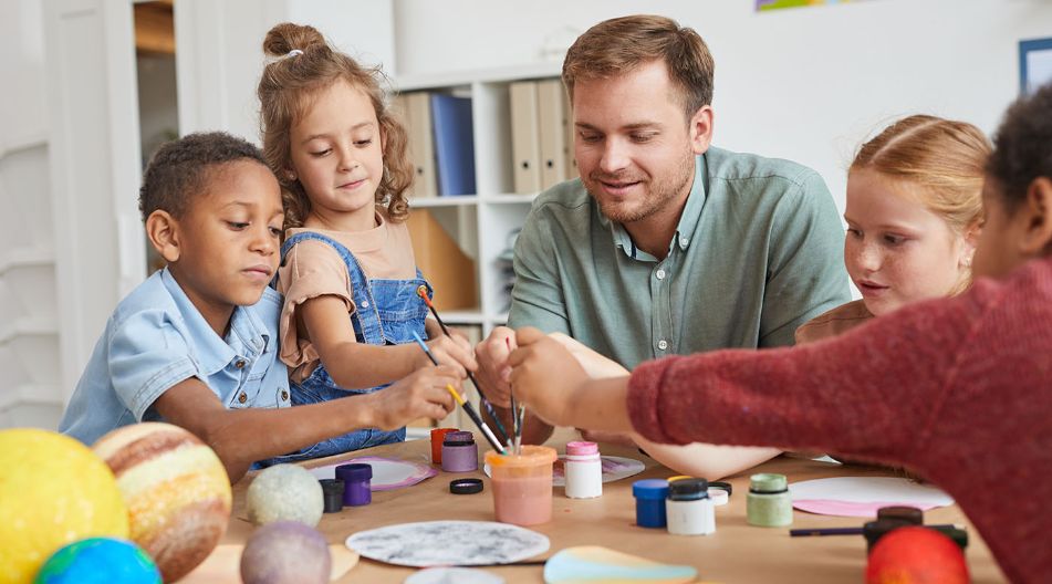 Ein Mann sitzt mit mehreren Kindern an einem Tisch und malt mit ihnen runde Blätter mit Tuschkastenfarbe an. Ein Mann sitzt mit mehreren Kindern an einem Tisch und malt mit ihnen runde Blätter mit Tuschkastenfarbe an.