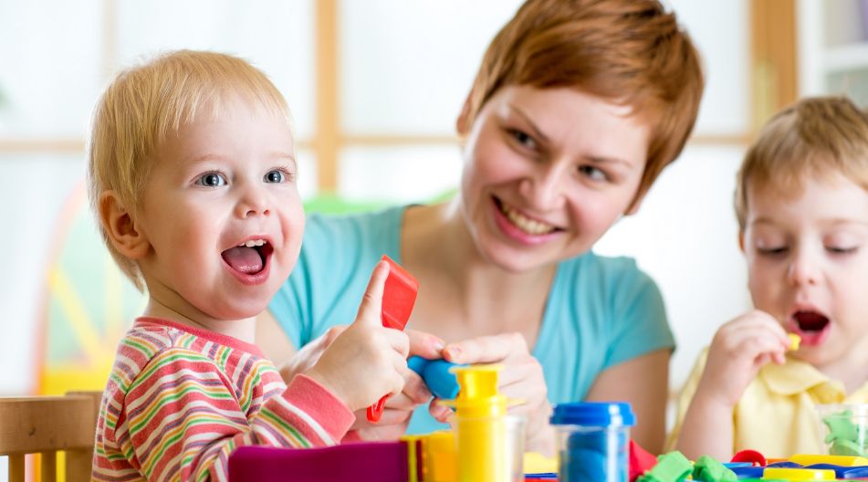 Eine Betreuerin spielt lachend mit zwei kleinen Kindern, die mit Knete basteln.