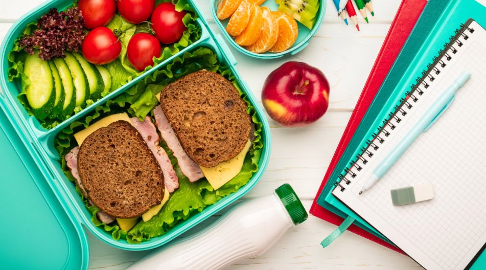 Ein geöffnetes Lunchbox-Set mit Vollkornbrotsandwiches, Tomaten, Gurkenscheiben, Blattsalat, Mandarinenstücken, Kiwi, und einem Apfel. Daneben liegen Schreibutensilien, Hefte und eine Trinkflasche – alles auf einem hellen Holztisch arrangiert. Ein geöffnetes Lunchbox-Set mit Vollkornbrotsandwiches, Tomaten, Gurkenscheiben, Blattsalat, Mandarinenstücken, Kiwi, und einem Apfel. Daneben liegen Schreibutensilien, Hefte und eine Trinkflasche – alles auf einem hellen Holztisch arrangiert.