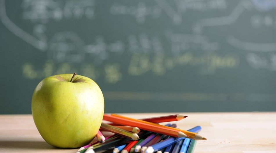 Ein grüner Apfel liegt neben einem Haufen bunter Bleistifte auf einem Holztisch. Im Hintergrund ist eine Schultafel mit unscharfen Zeichnungen und Formeln zu sehen. Ein grüner Apfel liegt neben einem Haufen bunter Bleistifte auf einem Holztisch. Im Hintergrund ist eine Schultafel mit unscharfen Zeichnungen und Formeln zu sehen.