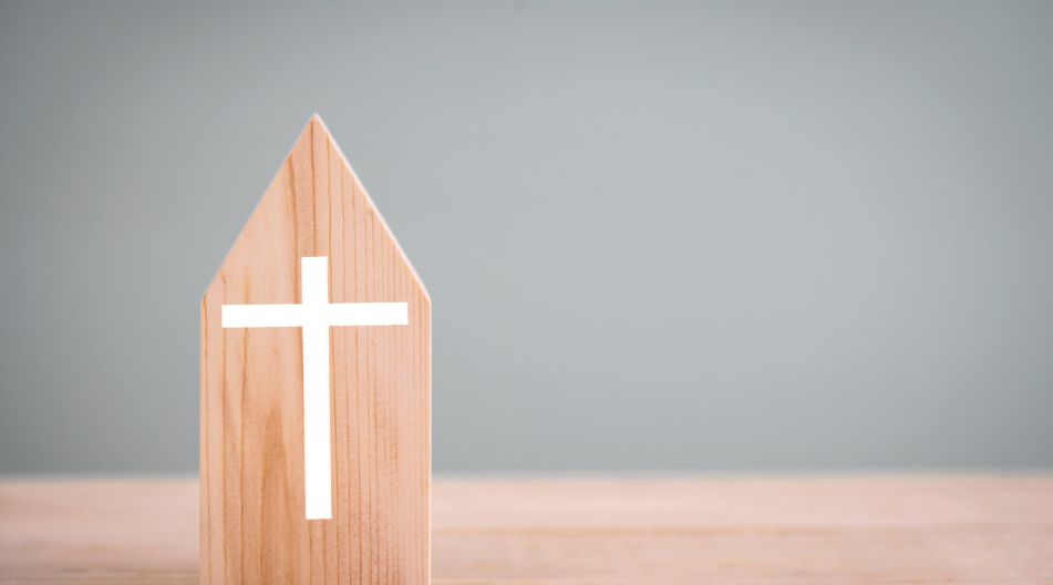 Ein Stück Holz geschnitzt in Form eines Hauses auf dem ein schlichtes weißen Kreuz ist. Das Stück Holz steht auf einem Tisch. Ein Stück Holz geschnitzt in Form eines Hauses auf dem ein schlichtes weißen Kreuz ist. Das Stück Holz steht auf einem Tisch.
