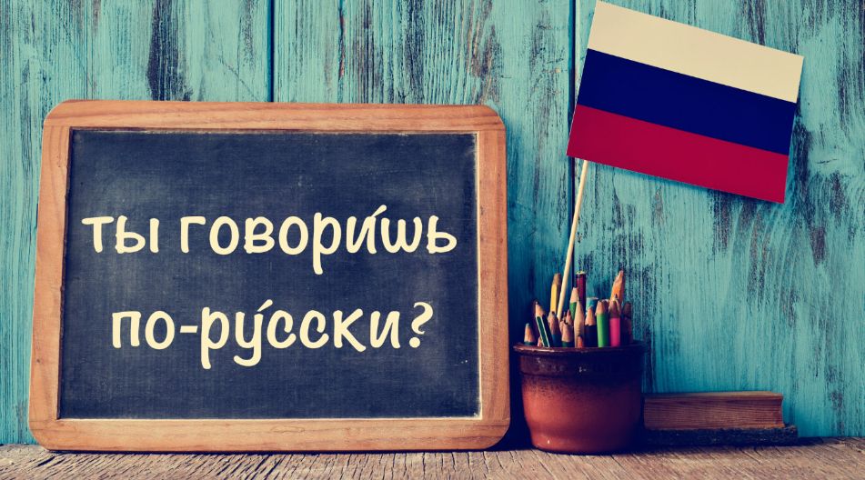 Eine Tafel mit der Aufschrift „Ты говоришь по-русски?“ vor einer blauen Holzwand, daneben ein Topf mit Buntstiften und eine kleine russische Flagge. Eine Tafel mit der Aufschrift „Ты говоришь по-русски?“ vor einer blauen Holzwand, daneben ein Topf mit Buntstiften und eine kleine russische Flagge.