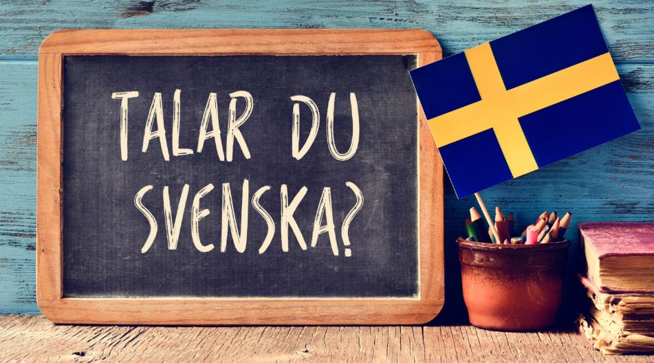 Eine Tafel mit der Aufschrift „Talar du svenska?“ vor einer blauen Holzwand, daneben ein Topf mit Buntstiften und eine kleine schwedische Flagge. Eine Tafel mit der Aufschrift „Talar du svenska?“ vor einer blauen Holzwand, daneben ein Topf mit Buntstiften und eine kleine schwedische Flagge.