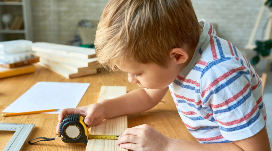 Ein Junge in einem gestreiften T-Shirt misst mit einem Maßband die Länge eines Holzbretts. Auf dem Tisch liegen Werkzeuge und weitere Holzstücke, die eine handwerkliche Tätigkeit andeuten. Ein Junge in einem gestreiften T-Shirt misst mit einem Maßband die Länge eines Holzbretts. Auf dem Tisch liegen Werkzeuge und weitere Holzstücke, die eine handwerkliche Tätigkeit andeuten.